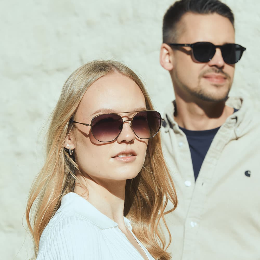 Blonde Frau mit dunkler Sonnenbrille und weißem, geripptem Poloshirt lehnt in hellem Sonnenlicht an einer Betonwand und berührt ihre Brille mit einer Hand.